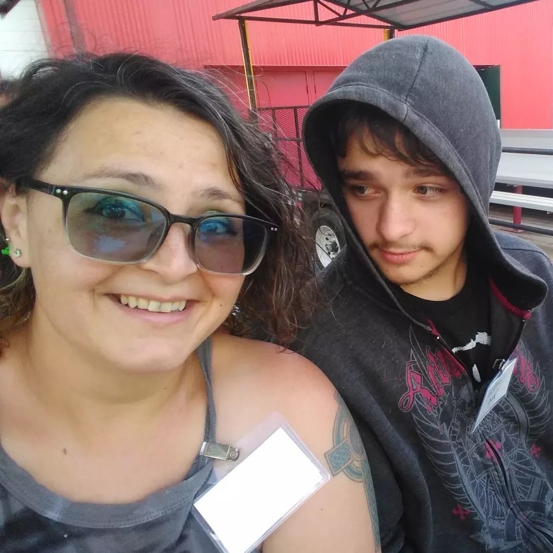 My son and I on the tour wagon.  Photo of woman in her late 30's with dark curly medium length hair, and a light olive skin tone.  She is wearing sunglasses and a grey tank top, a tattoo of a Celtic cross is partially visible on her left shoulder. She is looking at the camera. The son is teenaged with short medium brown hair and peach fuzz mustache. He wears a black shirt and dark grey hooded sweatshirt over his head. His gaze is on his mother.