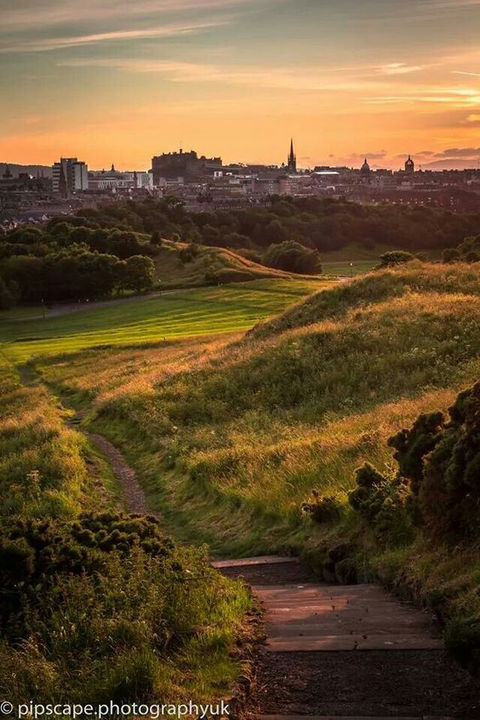 An Edinburgh skyline in the distance, against a setting sun. A park stretches across the foreground.