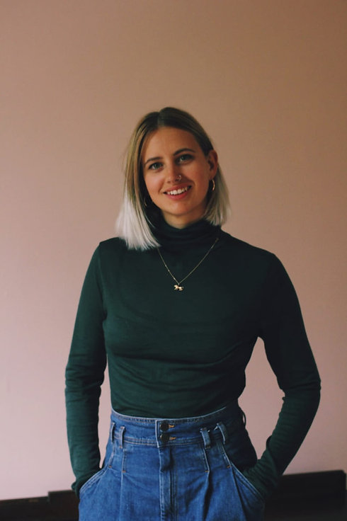 Leonie Rae Gasson: a white woman in her late twenties smiles at the camera. She has brown and blonde hair and is wearing a green poloneck, she stands against a soft pink background.