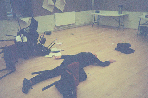 Tim lies face down on the floor, his outline looks like a dead body. He's surrounded by piled chairs and paper scattered across the floor.