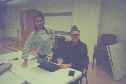 Mel and Tim look directly at the camera. Mel poses, pretending to be professional, Tim points a laptop, a beanie perched on his head.