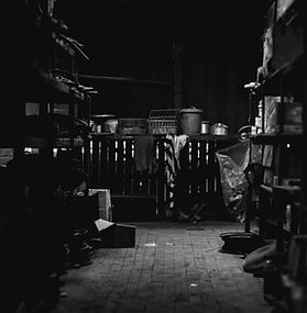 A dimly lit storage room with wooden floors and shelves lining both sides. Pots, containers, and household items sit at the far end of the room, illuminated by soft light filtering through gaps in the walls, while much of the space remains in deep shadow.