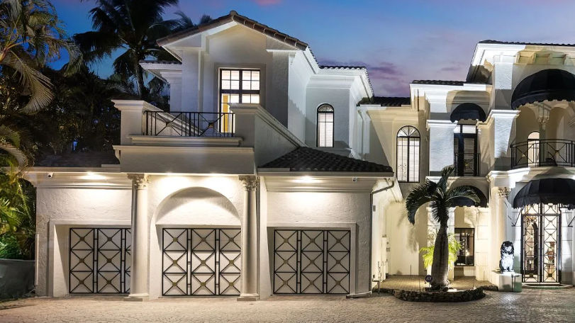 Elegant white mansion at dusk with arched windows, palm trees, and illuminated driveway. Clear sky with a hint of purple.