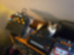 A cat has found herself behind a modular synthesizer which has two elektron synths and an arturia mini frieak on top.