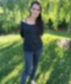 Junge Frau steht lächelnd im Gras vor grünem Baumbestand.