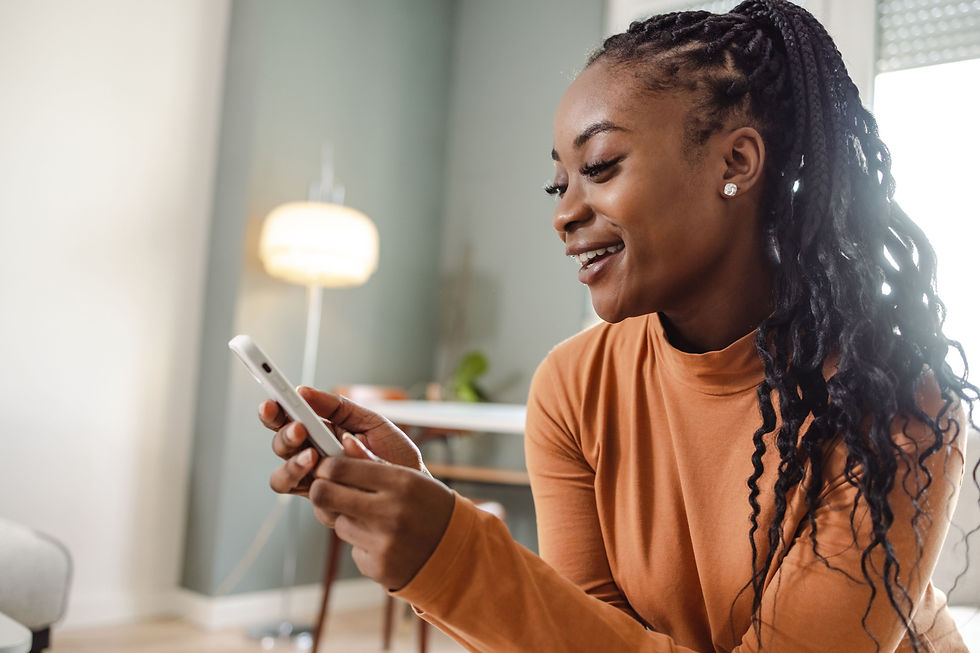 Smiling woman in an orange top looks at her smartphone in a bright home interior with a lamp and window behind her.