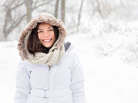 Smiling person in a white coat and fur-lined hood, standing in snowy outdoors with trees in the background. Snowflakes falling gently.