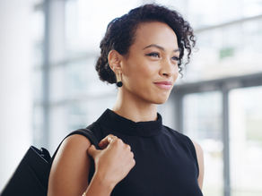 Smiling woman in black top holds bag strap, standing indoors with blurred windows in the background. She appears confident and content.