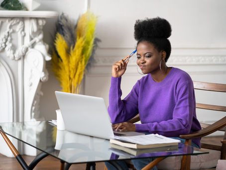 Woman in purple sweater works on laptop at glass table, holding pen thoughtfully. Background features white ornate mantel and yellow decor.