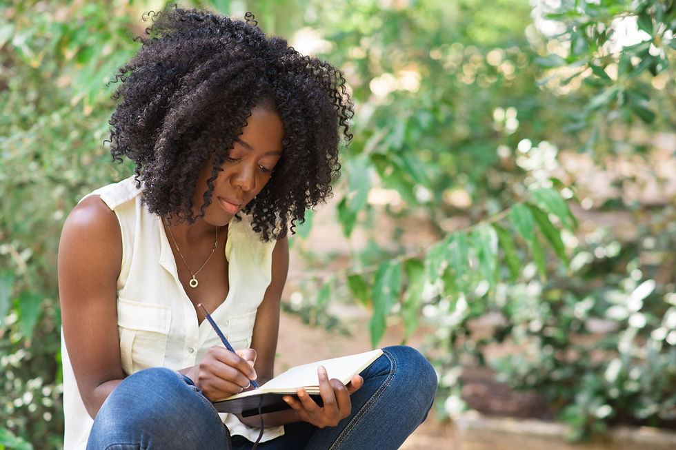 Woman with curly hair writing in a notebook outdoors. She's wearing a white top and jeans, surrounded by lush green foliage, focused and serene.
