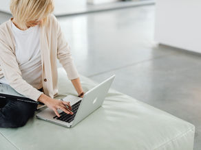 Person with blond hair sits on a light green couch, typing on a silver laptop. They wear a beige cardigan and hold a book with black cover.