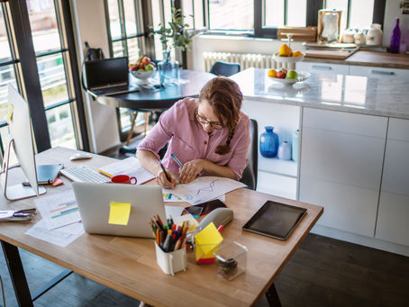 Woman in a pink shirt works at a desk in a bright home office, taking notes. A computer, tablet, and colorful fruit bowl are visible.