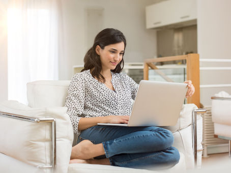 Woman in patterned shirt using a laptop, sitting on a white couch in a bright living room, smiling and relaxed. Soft natural light filters in.