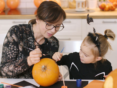 Woman and child in a kitchen painting a pumpkin. The child wears bat-themed headband. Orange and black colors dominate the decor.
