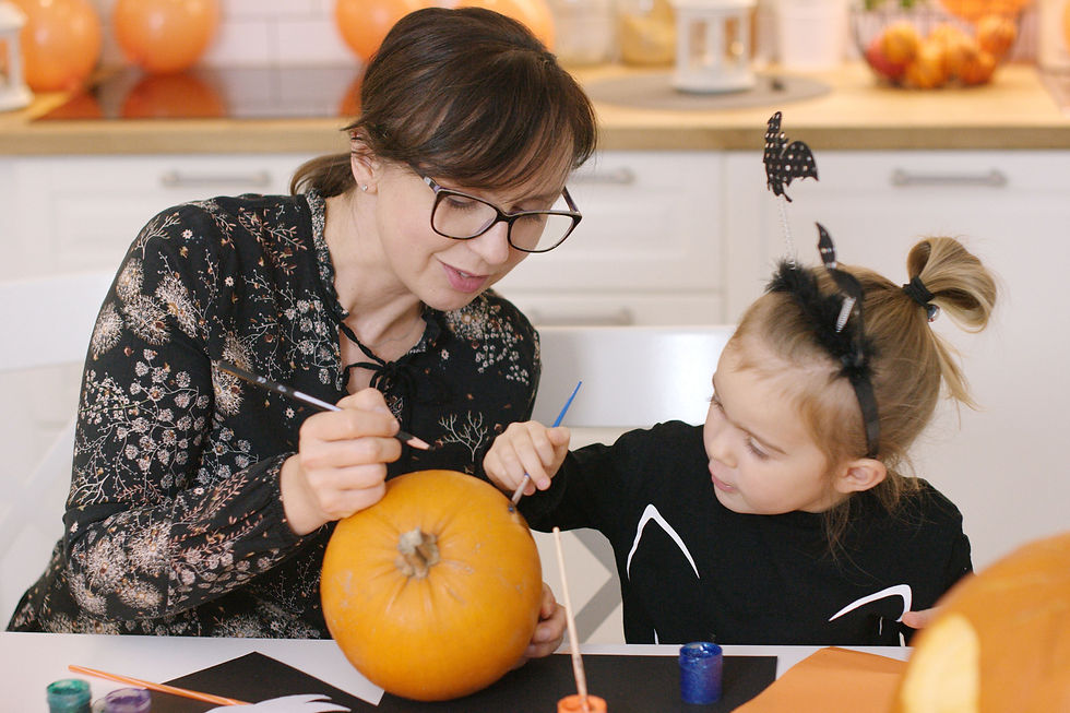 Woman and child in a kitchen painting a pumpkin. The child wears bat-themed headband. Orange and black colors dominate the decor.