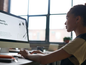 Woman working at a computer in a bright office. Screen shows "Identify your topic." Focused expression, windows in the background.