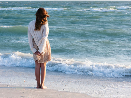 Woman in white sweater and beige skirt stands barefoot on a sandy beach, facing the ocean. Wind blows her hair, evoking a peaceful mood.