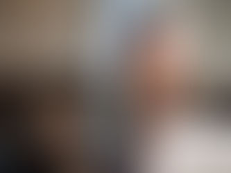 Smiling woman in a patterned jacket sits in a cafe with a laptop and coffee cup. Blurred background with warm tones enhances the cozy mood.