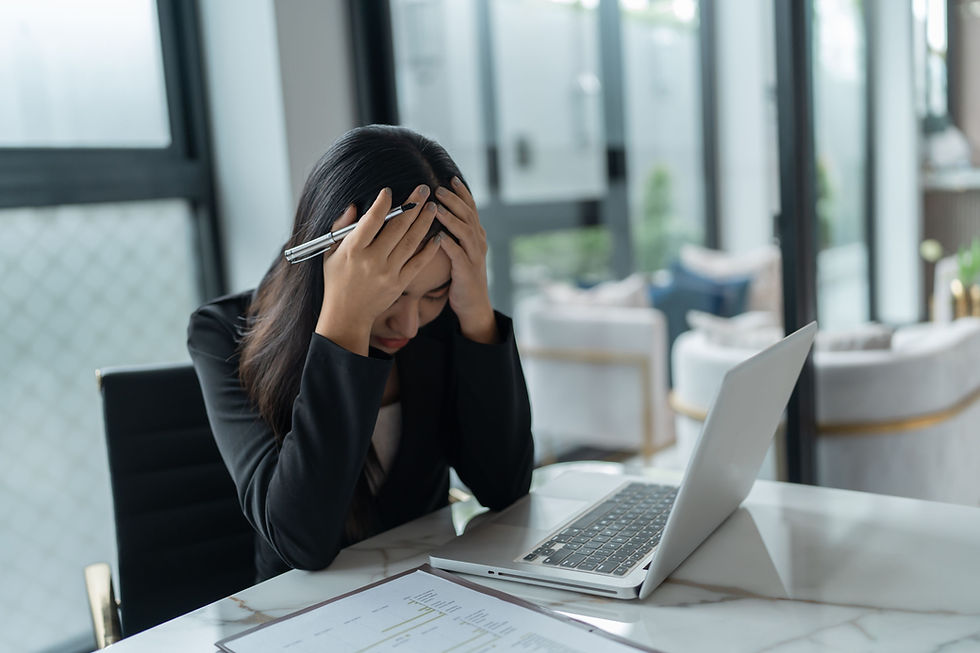 A person sits at a desk with a laptop, holding their head in frustration. Papers are scattered, and the office setting has large windows.