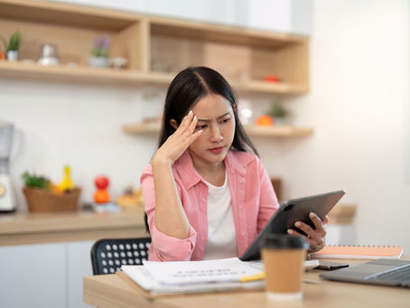 Woman in pink shirt, sitting at a kitchen table with a laptop and coffee, looks stressed while holding a tablet. Shelves with plants behind her.