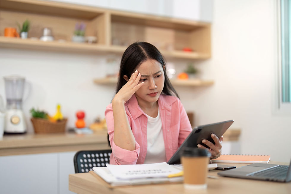 Woman in pink shirt, sitting at a kitchen table with a laptop and coffee, looks stressed while holding a tablet. Shelves with plants behind her.