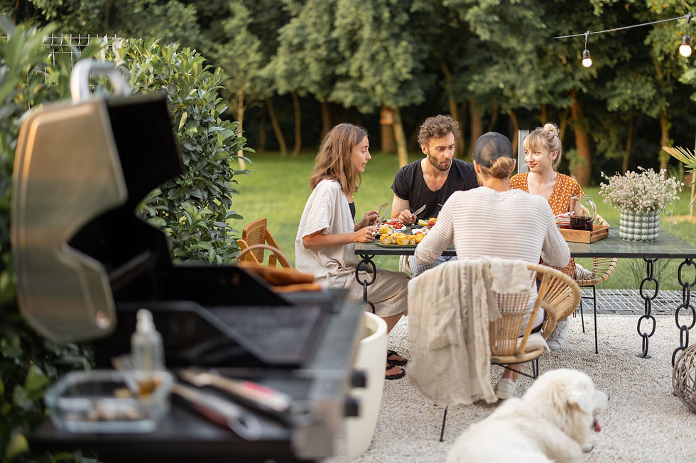 Four people enjoying a meal outdoors at a table, surrounded by greenery. A white dog lies nearby, and a grill is in the foreground.