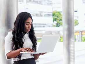 Woman in a white shirt using a laptop outdoors, focusing intently. Background shows blurred buildings, giving a professional vibe.