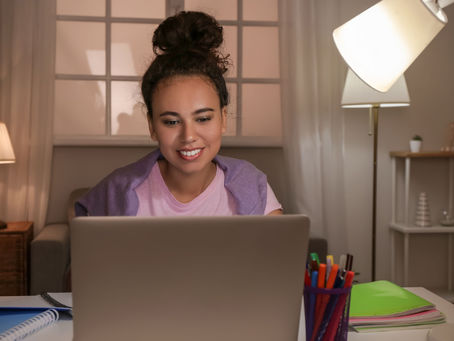 Woman smiling at a laptop in a cozy room with lamps and shelves. Notebook and colorful pens on the desk. Soft, warm lighting.