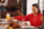 Woman in red sweater holds hands at festive table with roast, juice, and holiday decor. Warm, joyful atmosphere in cozy setting.
