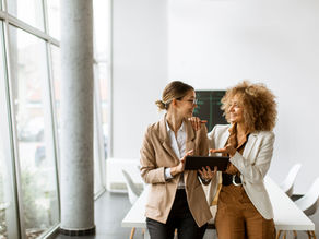 Two women in an office smile while looking at a tablet. Bright, modern setting with large windows and white furniture conveys positivity.