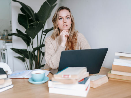 Woman in beige sweater sits at a table with a laptop, books, and a notebook, thinking. A green plant and soft light create a calm mood.