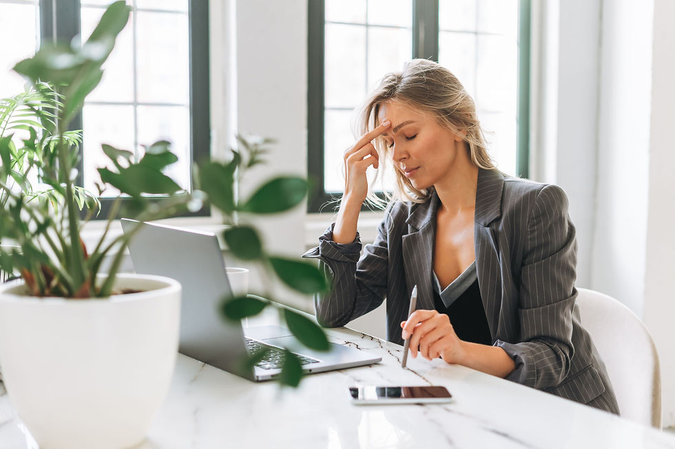 Woman in pinstripe suit sits at a table with a laptop, touching her forehead, looking stressed. Plants and a smartphone on the table.