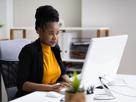 Woman in yellow shirt and black jacket working on a computer in a modern office. Potted plant on desk, shelves in background. Focused mood.