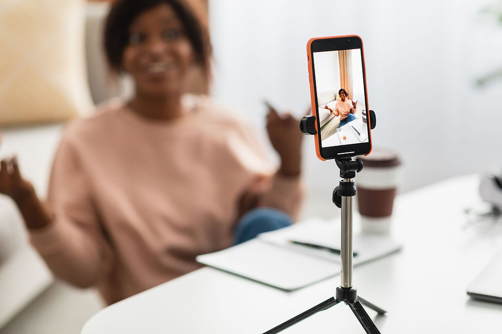 A person in a peach sweater is recorded by a phone on a tripod, indoors. Notebook and coffee cup are on the desk, bright setting.