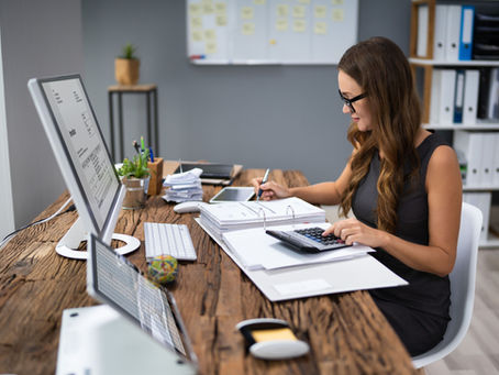 Woman in office working at a wooden desk, using a calculator and pen on documents. Computer screen displays an invoice. Shelves in background.
