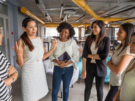 Women in an office setting engaged in lively conversation. One gestures animatedly while others listen and smile. Yellow pipes visible above.