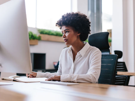Woman in a white blouse works at a desktop computer, writing in a notebook in a bright office, focused and calm.