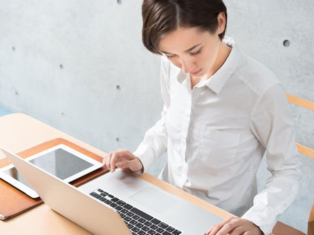 Woman in white shirt working on a laptop at a wooden desk, with a tablet beside her. Concrete wall in the background. Calm focus.