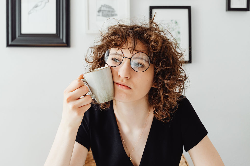 Curly-haired person in glasses holds a mug near face, sitting pensively. Black shirt, framed art on white wall in background.