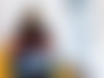 Woman in a cap works on a sticker-covered laptop in a bright room. Her expression is focused. She's seated on a yellow chair.