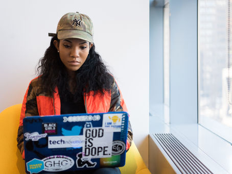 Woman in a cap works on a sticker-covered laptop in a bright room. Her expression is focused. She's seated on a yellow chair.