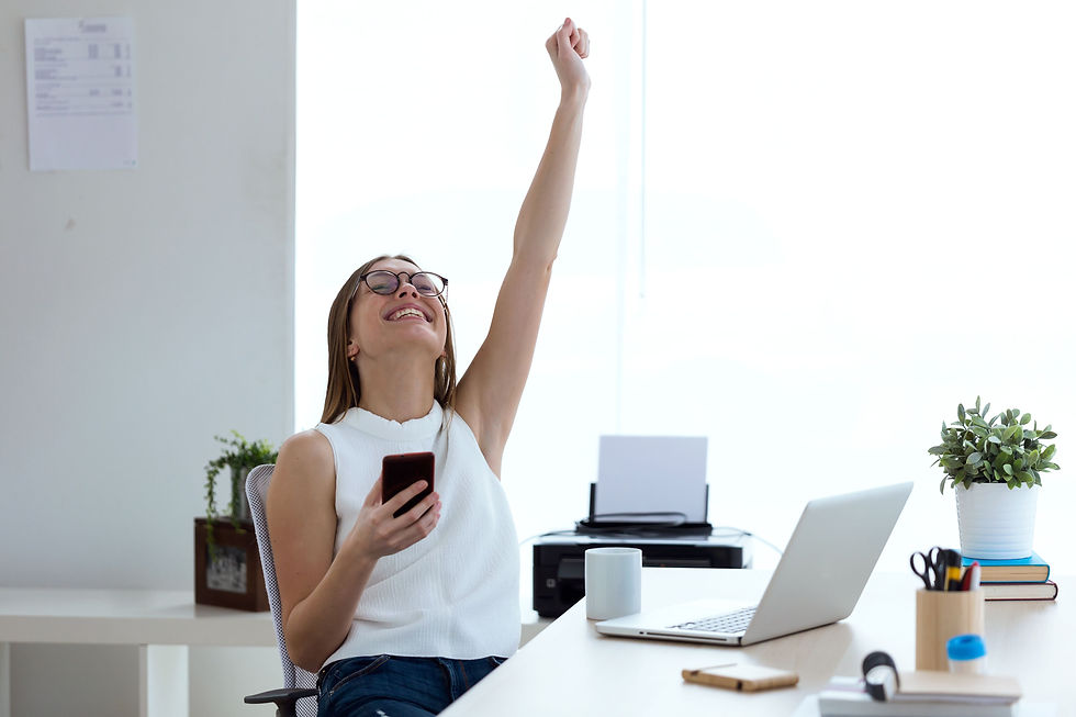Smiling woman in bright office raises one arm while holding phone, seated by laptop and plants.