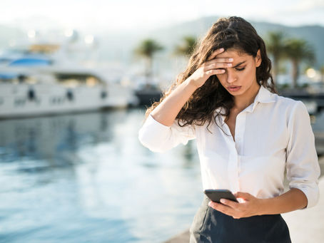 Woman in a white blouse looks worried while checking her phone. She's standing near a marina with yachts and palm trees in the background.