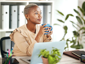 Woman with short hair holds a blue mug at a desk with a laptop in a bright office, looking thoughtful.