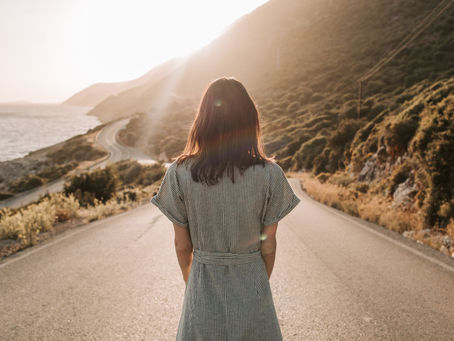 Woman in a striped dress stands on a sunlit road by the sea, with mountains in the background, evoking a serene and contemplative mood.