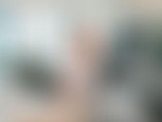 Smiling woman in a bright office holds glasses, types on laptop. Background features large windows, a potted plant, and empty chairs.