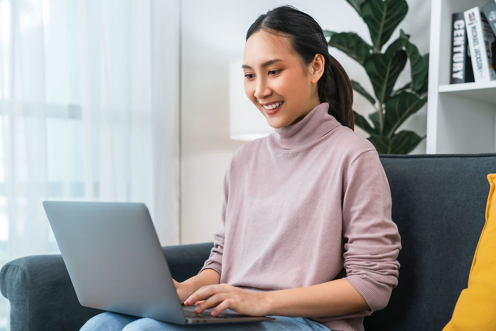 Smiling woman in a pink turtleneck uses a laptop on a sofa in a bright living room with plants and books.