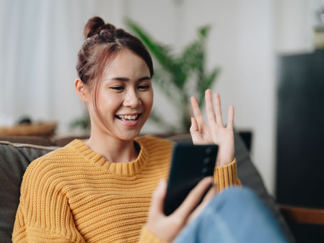 Smiling woman in yellow sweater waves at phone, sitting on a couch. Green plant in background. Warm, cheerful atmosphere.
