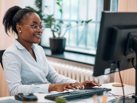 Woman in glasses typing on a keyboard in a modern office, smiling. Background has a large window and a potted plant. Bright and cheerful mood.