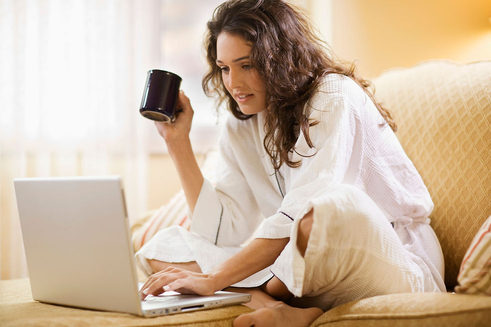 Woman in a white robe sits on a couch, typing on a laptop and holding a mug in a warm, cozy living room.